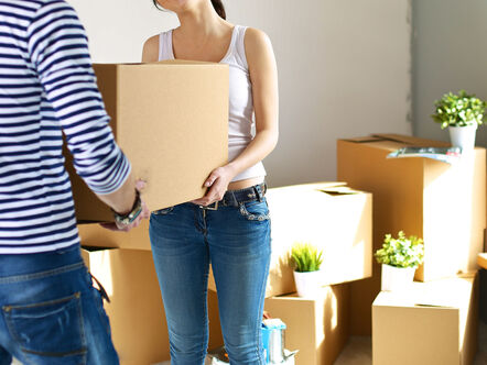 Two people carrying a cardboard box with other boxes in the background